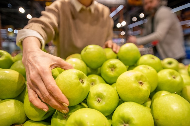 Mano del consumatore femminile maturo che prende la mela fresca di granny smith mentre fa una pausa il mucchio di frutta durante la visita al supermercato