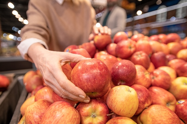 Mano della femmina matura cliente scegliendo mele rosse fresche sul display di frutta durante l'acquisto di prodotti alimentari con il marito in un supermercato