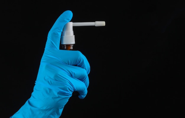 Hand in a medical glove holds a bottle of throat spray on a black wall.