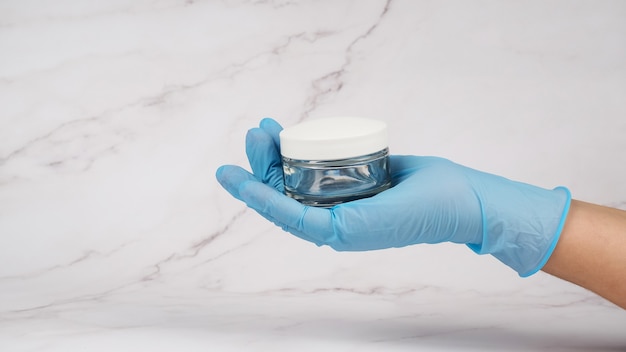 A hand in a medical glove holds a jar of cream