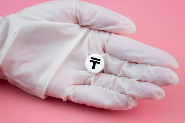 A hand in a medical glove holds a pill with a tenge sign on a pink background Expensive medicine in Kazakhstan Pharmaceutical business