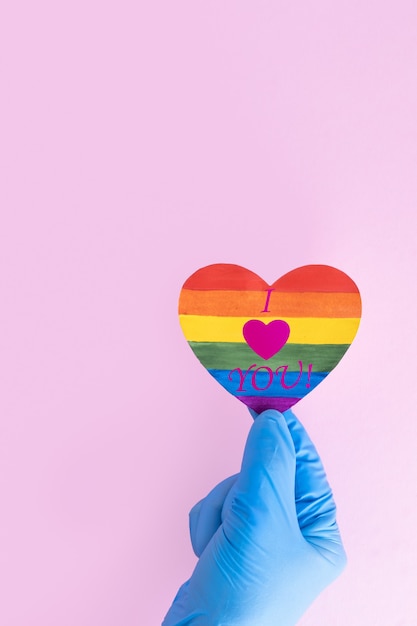 Una mano in un guanto protettivo medico tiene una forma di cuore di carta arcobaleno con la scritta i love you su uno sfondo rosa, cornice verticale, spazio copia. concetto di amore sicuro. concetto lgbt