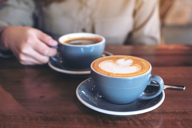 Hand met een blauwe kop warme zwarte koffie met een ander kopje koffie latte op houten tafel in café