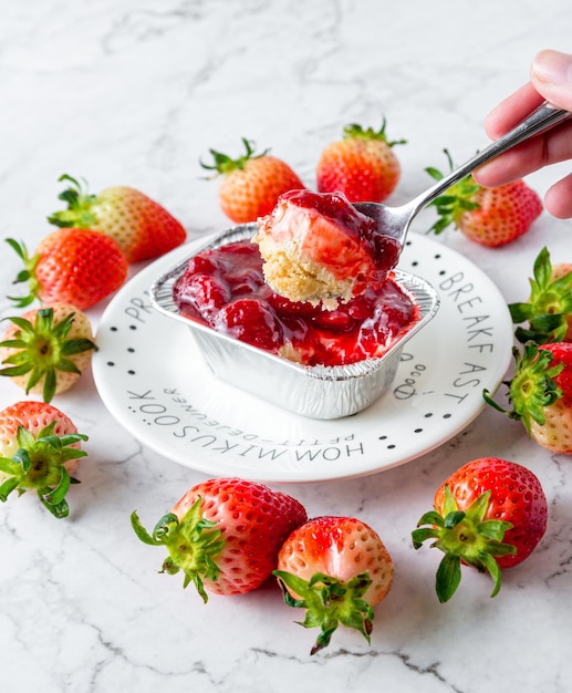 Hand met een lepel schepte de aardbeienkaastaart op Aardbeienkaastaart is verpakt in een vierkante aluminiumfolie in een witte plaat op een marmeren tafeltaart in café of bakkerij