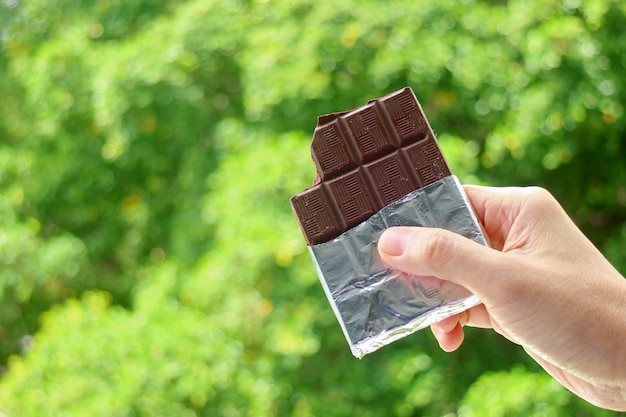 Hand met een reep pure chocolade in folieverpakking met wazig groen gebladerte op de achtergrond