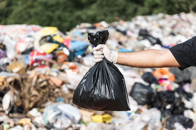 Hand met een zwarte vuilniszak voor een grote berg en een stapel afval. vuilnisplastic uit de industrie en de gemeenschap. wereld milieu dag.