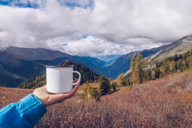 Hand met emaille witte mok mockup met bos en bergen vallei achtergrond Trekking merchandise en kampeerspullen Wildwood voorraad foto met wit metalen kop Rustiek product sjabloon