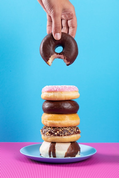 Hand met gebeten donut en stapel verschillende donuts op roze en blauwe achtergrond.