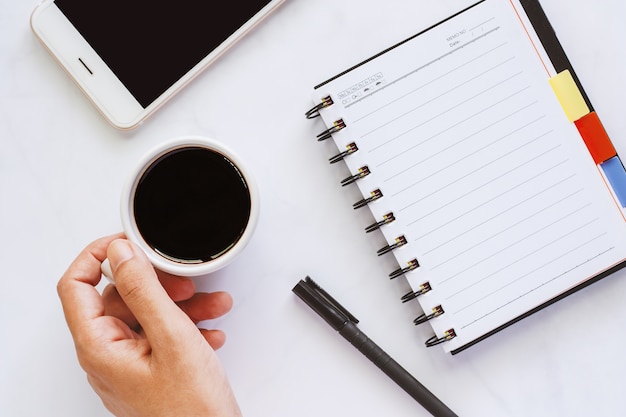 Hand met kopje koffie met spiraal notebook, pen en smartphone