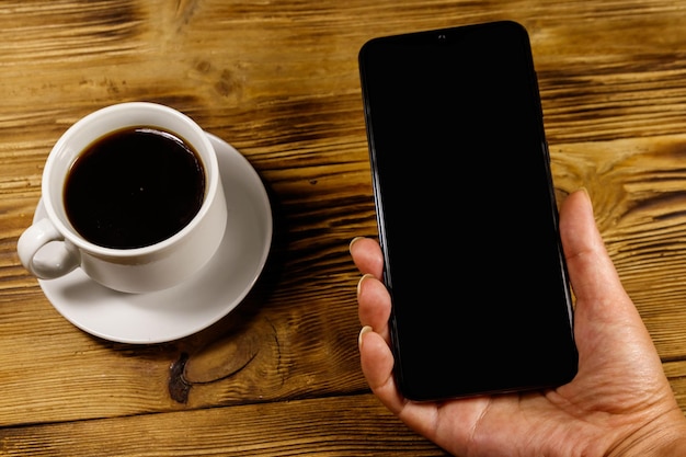 Hand met moderne smartphone en kopje koffie op een rustieke houten tafel