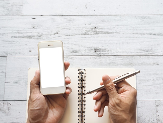 Hand met smartphone witte display en pen op blanco pagina notitieboekje op witte houten tafelblad weergave