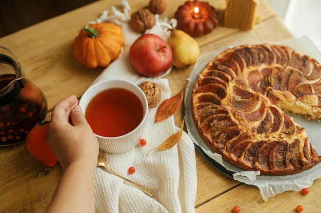 Hand met warme thee in stijlvolle beker op rustieke tafel met versgebakken appeltaart Herfst hygge