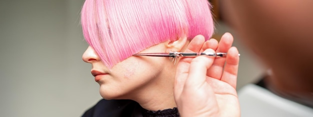Рука парикмахера стрижет короткие розовые волосы ножницами в парикмахерской крупным планом, вид сбоку