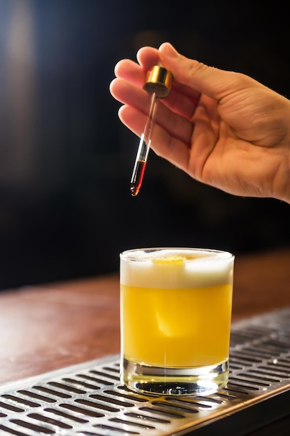 Рука бармена-мужчины держит пипетку и добавляет каплю жидкости в стакан с коктейлем на барной стойке
