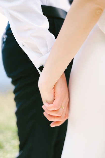 写真 白いシャツと黒いズボンを着た新郎の手は、白いドレスを着た花嫁の手を握っています。閉じる。高品質の写真