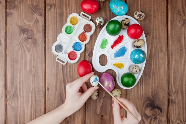 Hand painted Easter eggs, paints and brushes on wooden background. Preparation for the holiday. Girls hands draw a pattern