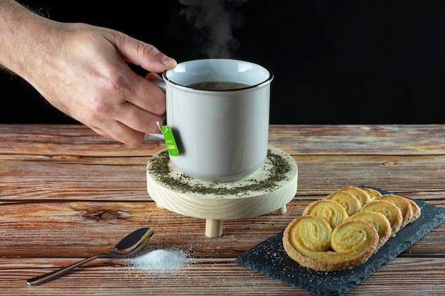 Photo hand of a person holding a cup of hot tea ready to drink on a wooden base accompanied by cookies sugar sprinkled on the table
