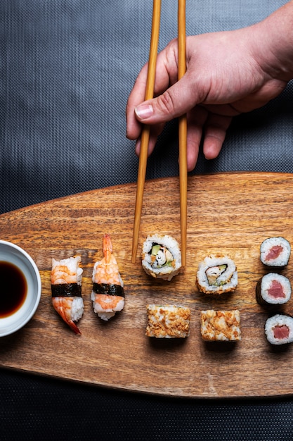 рука, собирание суши Ролл с деревянной доски. Состав суши Маки, Нигири и Калифорния Ролл с палочками для еды и соевым соусом.