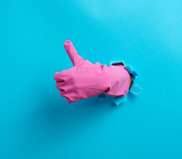 Hand in a pink household rubber glove sticks out of the torn hole and shows a gesture okay thumb up blue background