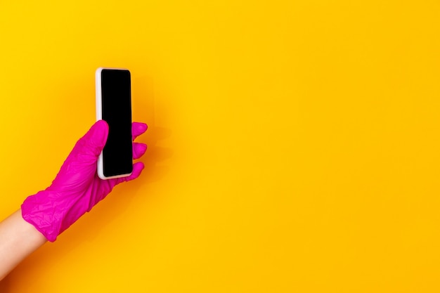 Hand in pink protective rubber glove with smartphone isolated on yellow studio background with copyspace. Gesturing, holding, presenting things. Negative space for your advertising. Showing, pointing.