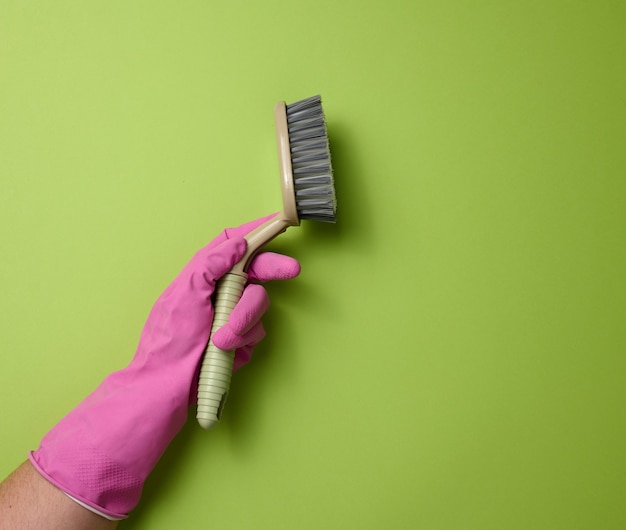Рука в розовой резиновой перчатке держит пластиковую щетку для чистки на зеленом фоне