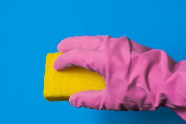 A hand in a pink rubber glove holds a yellow sponge. Blue background.