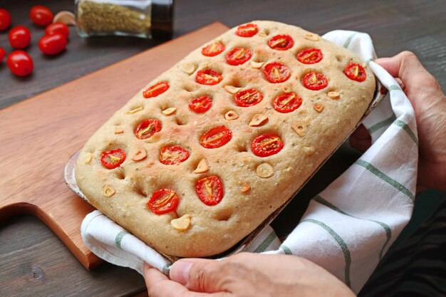 Hand Placing a Pan of Freshly Baked Italian Tomato and Garlic Focaccia Bread on Wooden Breadboard