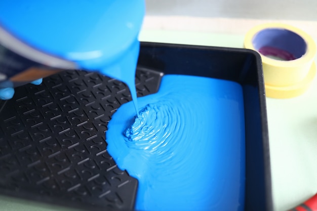 Hand pour blue paint from can into black plastic tray.