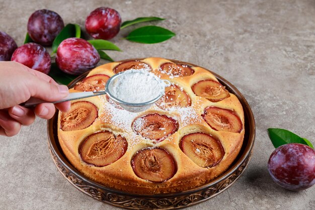 Hand pouring powdered sugar on red plum pie.