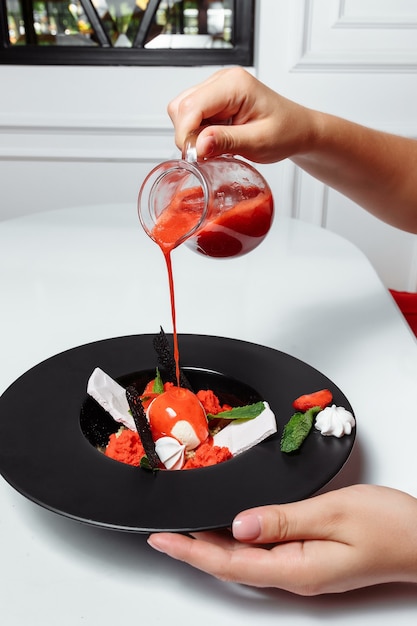 Hand pouring strawberry sauce to ice cream with fresh strawberries and decorated with mint.