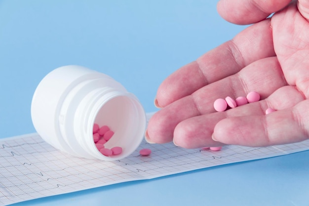 A hand pours a handful of pink pills from a white jar onto an electrocardiogram of the heart on a blue background The concept of a healthy lifestyle and timely medical examination