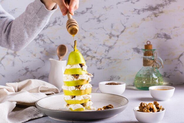 A hand pours honey over a pear with ricotta and walnuts in layers on a plate Healthy food