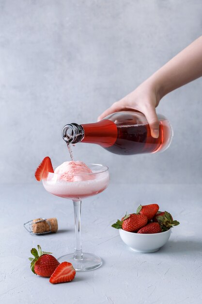 Hand pours pink champagne into a glass with strawberry ice cream on a gray surface. The concept of delicious drinks. Copy space.