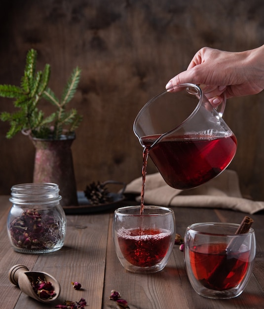 A hand pours red rose tea from a glass teapot into clear cups on a brown wooden table. front view
