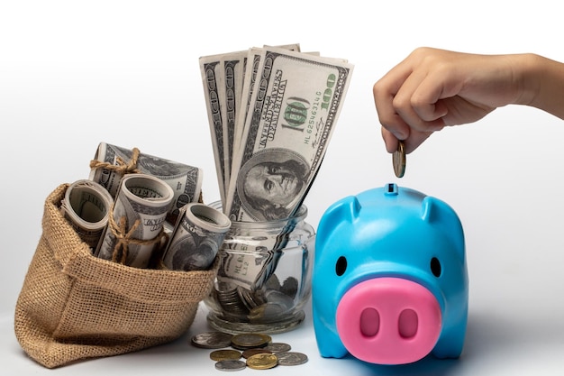 A hand put a coin into a piggy bank with a lot of coins and banknotes nearby.