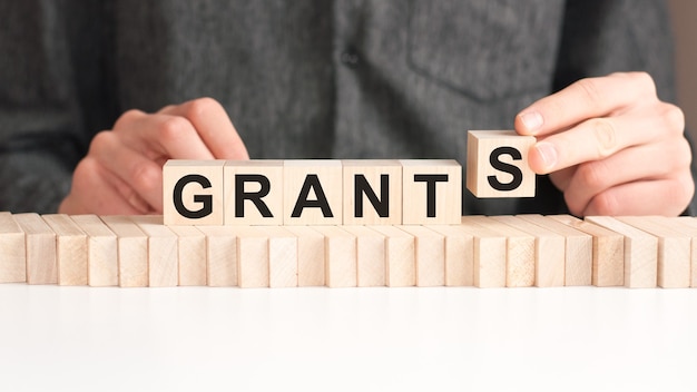 The hand puts a wooden cube with the letter S from the word GRANTS. The word is written on wooden cubes standing on the white surface of the table.