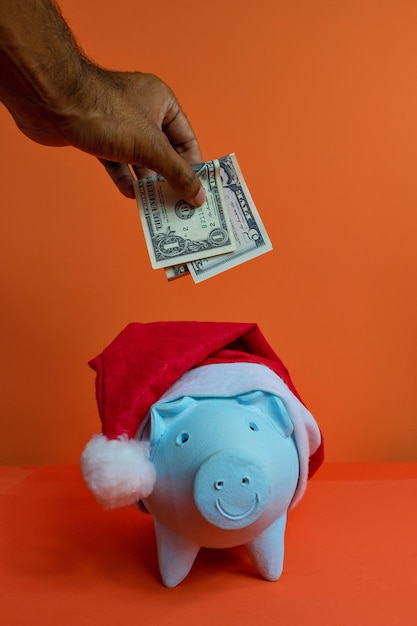 Hand putting dollars money in piggy bank with christmas Santa hat isolated on orange background. Concept image.