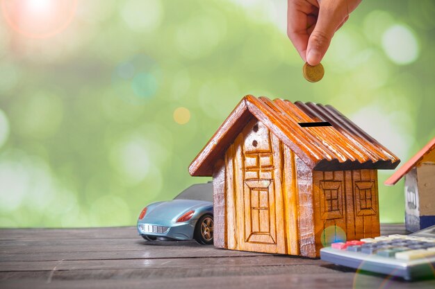 Hand putting golden coins in money wooden house model. Concept of savings plan for housing