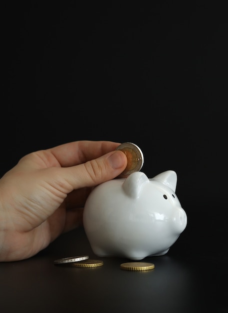 Hand putting money coin into piggy bank for saving money. Wealth, budget, investment, finance concept. Money box, piggybank on the black background.