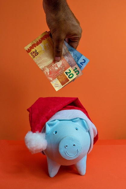 Hand putting real brazilian money in piggy bank with christmas santa claus hat isolated on orange background. Concept image.