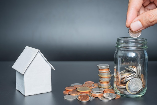 Hand putting thailand coins in jar with heap coins on black
background with house model