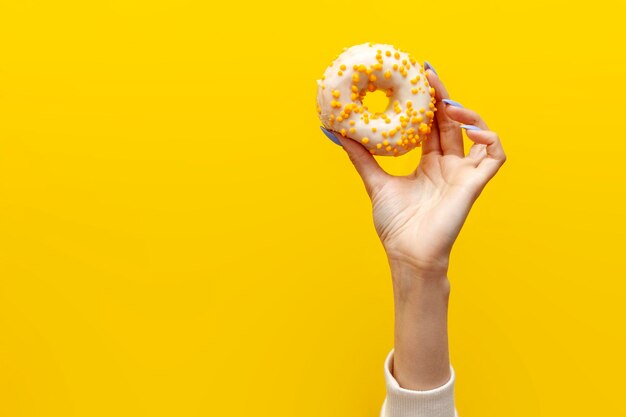 Hand raises and holds one sweet donut in white icing on yellow isolated background