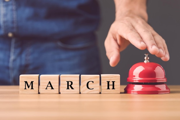 Hand ringing the red call bell and wooden blocks write word MARCH arranged on wooden desk with grey space wall background Start for new idea in March concept