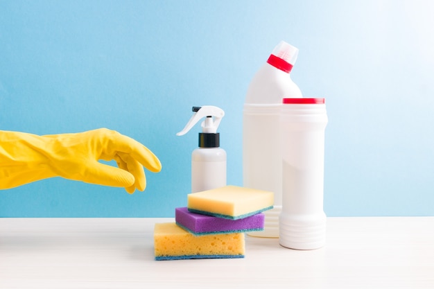 Hand in rubber glove reaches for a cleaning sponge, white bottles without labels with detergents and spray on the table