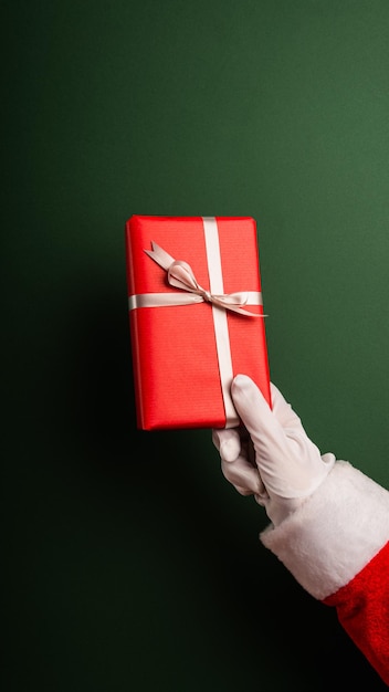 Hand of santa claus gives a red wrapped box with ribbon on a\
green background with copy space.