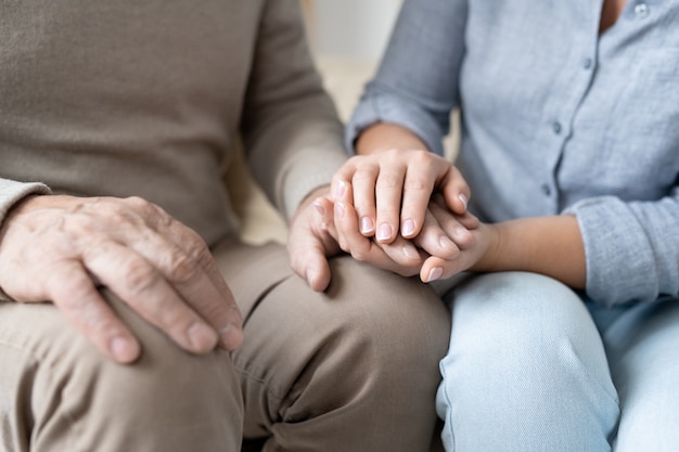 Hand of senior man in casualwear between those of his affectionate young daughter while both sitting close to each other