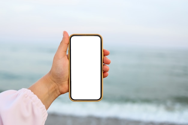 Hand showing a blank smart phone on the beach with the sea in the background. White screen mock up