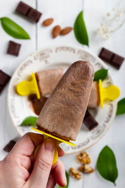 Hand showing in the foreground a delicious and refreshing chocolate ice cream lolly on a white wooden table covered with pieces of chocolate, nuts, almonds and leaves from nature