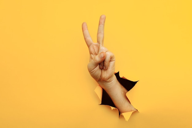 Photo hand showing peace gesture through torn yellow paper background victory v sign