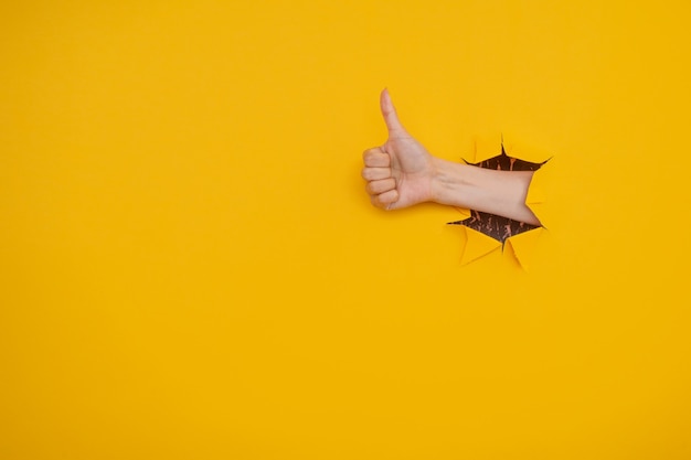 Hand showing a thumb up sign through a ripped hole in yellow paper wall. Well done, good job concept